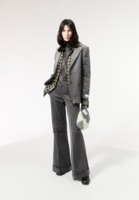 Grijze pak met een gestructureerde blazer, uitlopende broek, gelaagd met een groene en zwarte geruite shirt. Accessoires omvatten een moderne gebreide tas.