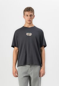 Young man wearing dark gray oversized T-shirt with colorful "Wild Soul" text on chest and light gray pants, standing against white background.