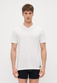 Weißes Baumwoll-T-Shirt mit kurzen Ärmeln und V-Ausschnitt, gerader Schnitt und einem schwarzen Logo-Akzent nahe dem Saum. Glatte Textur, leichtes Design.