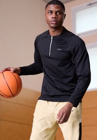 Fekete, hosszú ujjú sportos felső, negyed cipzárral és logóval, világos sárga tréningnadrággal, fekete kiegészítővel párosítva. A játékos egy narancssárga kosárlabdát tart a kezében.