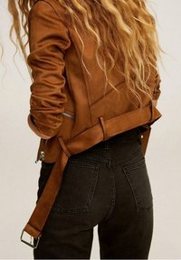 Brązowa zamszowa kurtka o dopasowanym kroju, z zamykanymi na zamek kieszeniami i pasek z tyłu, w zestawie z czarnymi dżinsami.