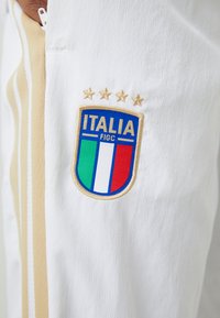 Λευκή αθλητική ενδυμασία που διαθέτει το επίσημο σήμα της ποδοσφαιρικής ομάδας της Ιταλίας με τέσσερα χρυσά αστέρια επάνω και μια κατακ�όρυφη ρίγα σε πράσινο, λευκό και κόκκινο χρώμα.