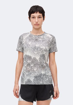 Γυναίκα με κοντά σκούρα μαλλιά που φοράει λευκό μπλουζάκι με μαύρα αφηρημένα σύννεφα και μαύρο αθλητικό σορτς.