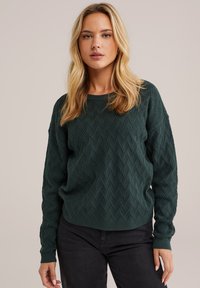 Pull en tricot vert foncé avec un motif en diamant texturé, col rond et manches longues, associé à un pantalon noir.