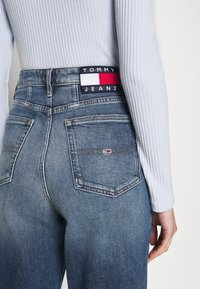 Jeans i medelblå denim med hög midja, prydda med en logotyppatch i rött, vitt och marinblått. Fem fickor och sliten textur på baksidan.
