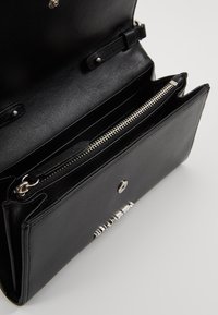 Portafoglio in pelle nera con scomparto con zip, chiusura a bottone e slot per carte interno. Finitura liscia con dettagli in metallo argentato.