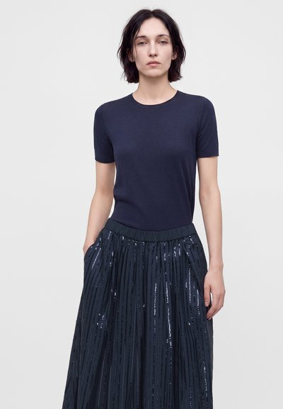 Femme aux cheveux courts et foncés portant un haut uni bleu marine à manches courtes et une jupe plissée foncée avec des bandes verticales de sequins, debout devant un fond uni.