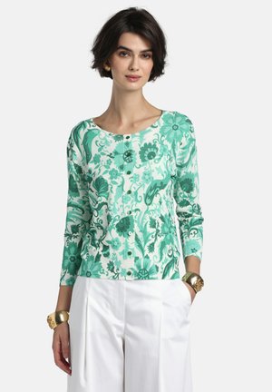 Žena oblečená v zelenom kvetovanom kardigáne na gombíky s bielymi nohavicami, zlatými náramkami a zlatými náušnicami, stojaca pred obyčajným pozadím.