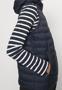 Henkilö, jolla on yllään tummansininen tilkkutäkki, ilman hihoja oleva liivi musta-valkoisten raidallisten pitkähihaisten paidan päällä, käsi liivin taskussa, yksinkertaista taustaa vasten.