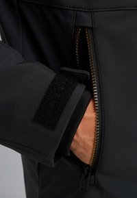 Manga de chaqueta negra aislante con puño de velcro, detalle de cremallera dorada y textura suave. Vista en primer plano de la mano y el cierre.