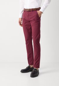 Chinos in cotone bordeaux con tasche laterali inclinate, una cintura in pelle marrone e mocassini neri. Texture liscia, vestibilità sartoriale e cuciture visibili.