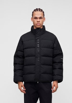Joven con cabello trenzado que lleva una chaqueta acolchada negra con cuello alto y texto de logo en el frente y la manga.