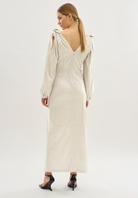 Una donna con una coda di cavallo bionda indossa un lungo vestito beige con dettagli annodati sulle spalle e schiena scoperta, abbinato a sandali neri con tacco alto.