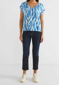 Top corto a maniche corte blu e bianco con motivo, abbinato a jeans di denim scuri e sneaker chiare. Il top presenta un collo a barco.