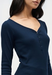 Femme portant un haut henley bleu foncé à manches longues avec un col en V et quatre boutons, présentée des épaules à la taille sur un fond uni.