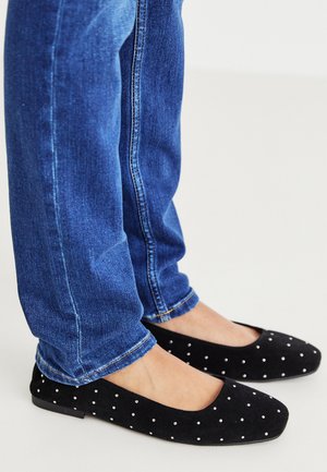 Ballerine in suede nero con pois bianchi, caratterizzate da una punta arrotondata e suola piatta, abbinate a jeans in denim blu.