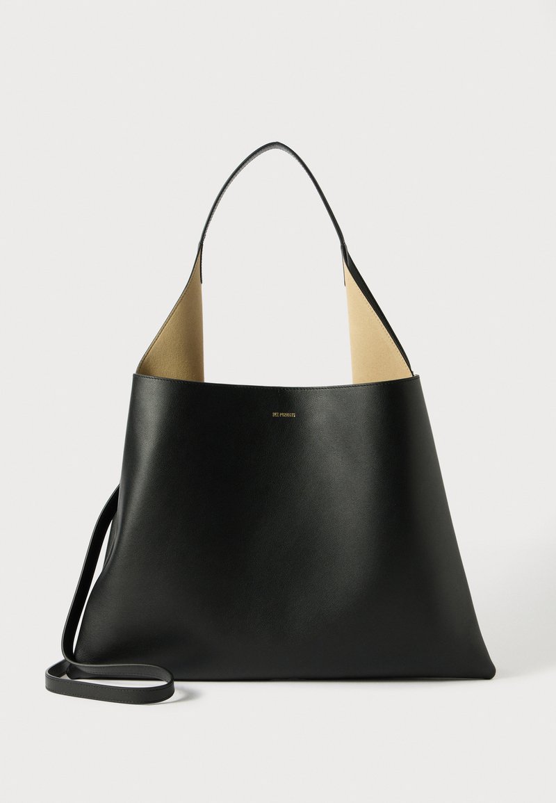 Borsa tote in pelle nera con un design spazioso e angolare. Presenta un interno in contrasto di color cammello e una sottile tracolla. Branding minimalista visibile.