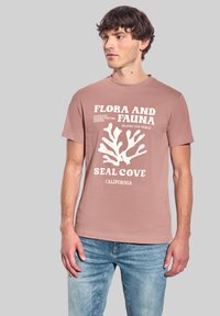 Ljusrosa bomulls t-shirt med en vit korallgrafik och texten "Flora och Fauna, Seal Cove, Kalifornien." Standard passform.