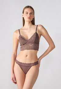 Smeđi čipkasti bralette s tankim trakama i valovitim rubovima, uparen s odgovarajućim tangama s čipkastim rubovima. Glatka tekstura, detalj cvjetnog uzorka.