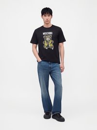 Νέος άνδρας που φοράει μαύρο t-shirt Moschino με γραφικό αρκουδάκι, μπλε τζιν και μαύρα αθλητικά, στέκεται μπροστά σε λευκό φόντο.
