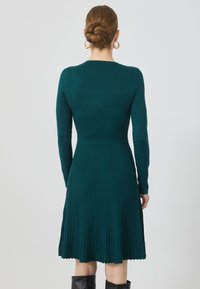 Rochie din tricot ribbed, cu mâneci lungi, de culoare teal, cu un corset ajustat care se transformă într-o fustă evazată cu margine în valuri. Design minimalist, fără modele.