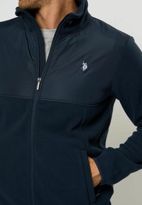 Giacca in pile navy con zip, caratterizzata da una parte superiore liscia e una texture morbida, tasche laterali e logo ricamato sul lato sinistro del petto.