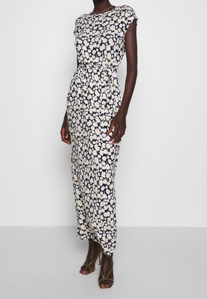 Femme portant une robe noire sans manches, longueur cheville, avec des motifs de marguerites blanches et jaunes, et des sandales à talons hauts vert olive.