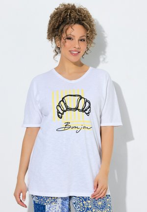 Kobieta z kręconymi włosami, ubrana w białą koszulkę z grafiką rogalika i napisem „Bonjour”, w parze z niebieskimi wzorzystymi spodniami, stojąca przy białej ścianie.