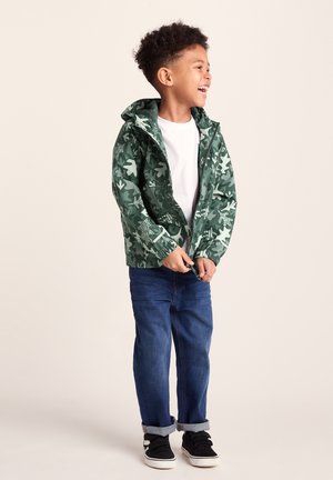 Young boy smiling and zipping a green camouflage jacket, wearing blue jeans and black sneakers against a neutral background.