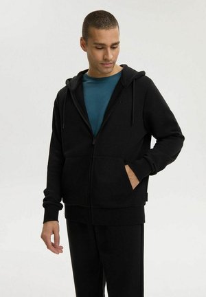 Mężczyzna ubrany w czarną bluzę z suwakiem i spodnie, pod którymi ma turkusową koszulkę, stoi z jedną ręką w kieszeni, patrząc w dół.