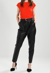 Mujer con una blusa roja sin mangas, pantalones de cuero negros de cintura alta con cinturón y zapatos de tacón alto negros, de pie con las manos en los bolsillos.