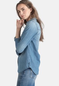 Chemise en denim bleu clair à manches longues, avec des poignets à boutons, un ourlet arrondi et des fentes sur les côtés, associée à un jean en denim foncé. Texture lisse.