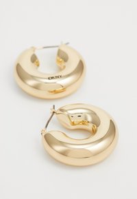 Pendientes de aro de oro con una superficie lisa y pulida. Forma redondeada en media luna que presenta una bisagra delgada de plata para el cierre. Marca DKNY visible.