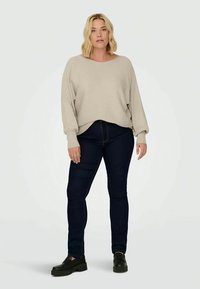 Blondýnka stojí oblečená v béžovém ribbovaném svetru, tmavě modrých skinny džínách a černých platformových botách na jednobarevném pozadí.