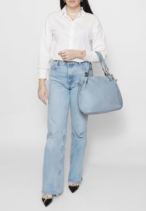 Femme portant une chemise blanche à boutons et un jean bleu clair, tenant un grand sac à main bleu clair, debout devant un fond blanc uni.