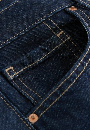 Detailný záber na tmavomodré džínsy s vreckom, na ktorom sú viditeľné švy a na hornom rohu je medený nit.