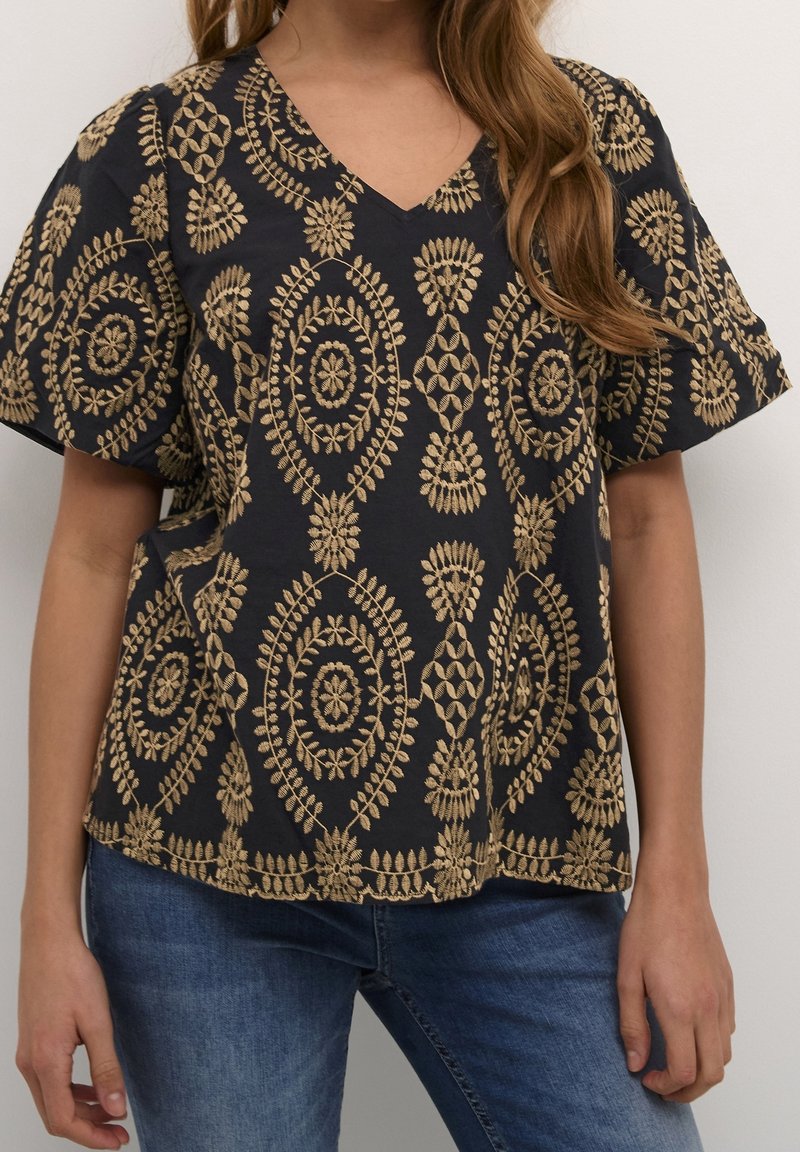 Blouse noire à manches courtes avec broderie florale beige portée avec un jean bleu par une personne aux longs cheveux châtain clair.