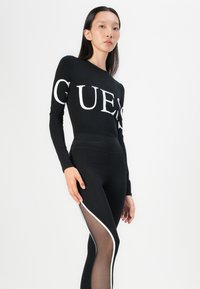 Črn dolg rokav vrhnji del z velikim belim logotipom "GUESS", v kombinaciji s črnimi pajkicami z prosojnimi stranskimi paneli in belim obrobo. Gladka tekstura materiala.