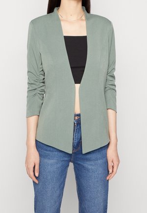Femme portant un blazer vert clair à manches plissées sur un crop top noir et un jean bleu, debout devant un fond uni.
