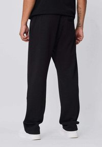 Pantalon de survêtement noir en tissu lisse, avec une coupe décontractée, une taille élastique et des ourlets côtelés ; associé à des baskets blanches.