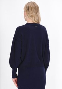 Granatowy sweter o luźnym kroju, szerokich rękawach, ściągaczach przy mankietach oraz małym metalicznym akcentem z tyłu przy karku. Miękka tekstura.