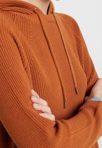 Oranssi neulottu huppari teksturoidulla viimeistelyllä, jossa on kiristysnyörillä varustettu huppu ja metalliset nyörinpäät. Hihat ovat hieman väljät.