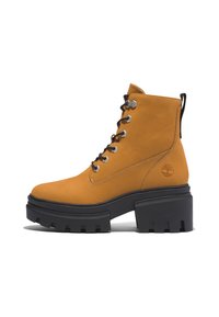 Stivaletto in pelle gialla con una robusta suola in gomma nera, occhielli argentati, lacci neri e un logo sul lato. Texture liscia e design resistente.