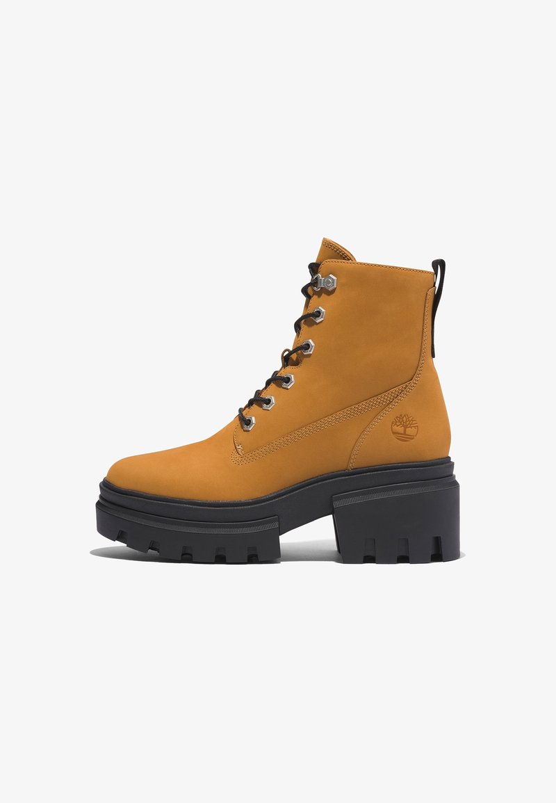 Stivaletto in pelle gialla con una robusta suola in gomma nera, occhielli argentati, lacci neri e un logo sul lato. Texture liscia e design resistente.