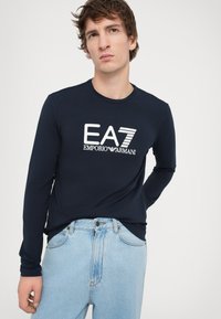 Marineblå langermet t-skjorte laget av myk bomull med hvit EA7 og Emporio Armani logo trykt på forsiden, sammen med lyseblå denimjeans.