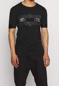 Schwarzes T-Shirt mit kurzen Ärmeln und Rundhalsausschnitt, das ein strukturiertes Paneel mit geometrischem Muster und ein zentrales Logo in kontrastierendem Gold aufweist.