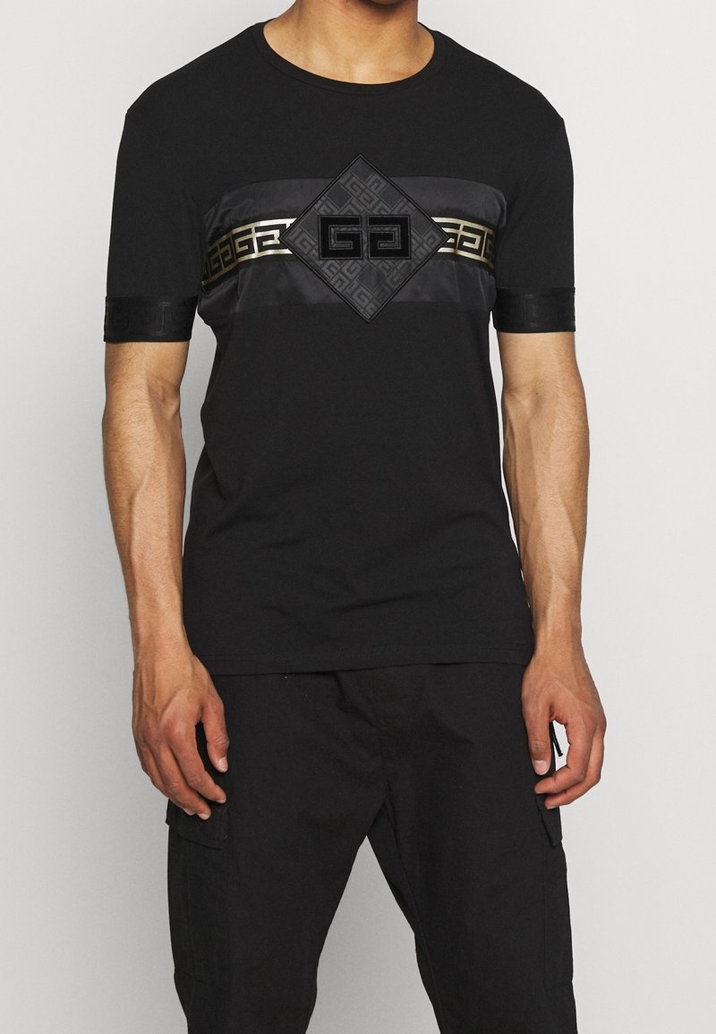 Schwarzes T-Shirt mit kurzen Ärmeln und Rundhalsausschnitt, das ein strukturiertes Paneel mit geometrischem Muster und ein zentrales Logo in kontrastierendem Gold aufweist.