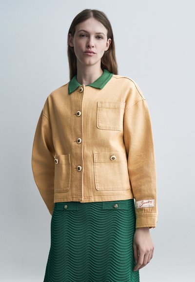Jeune femme portant une veste beige boutonnée avec des poches avant et un col vert, associée à une jupe verte texturée.