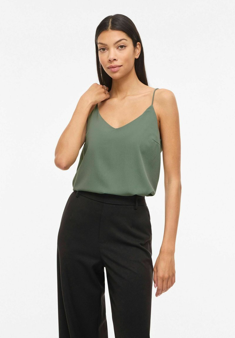 Kvinde med langt mørkt hår iført en ærmeløs grøn top og sorte bukser med højtaljet talje, poserer foran en hvid baggrund.