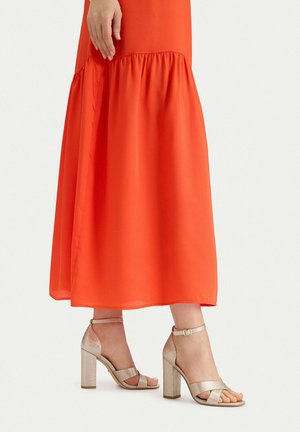 Frau trägt einen langen, fließenden orangen Rock und metallic goldene High-Heel-Sandalen mit Knöchelriemen, steht vor einem schlichten Hintergrund.